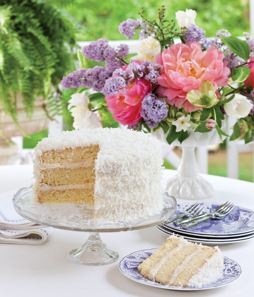 Layered coconut cake on a stand in front of pink and purple flowers