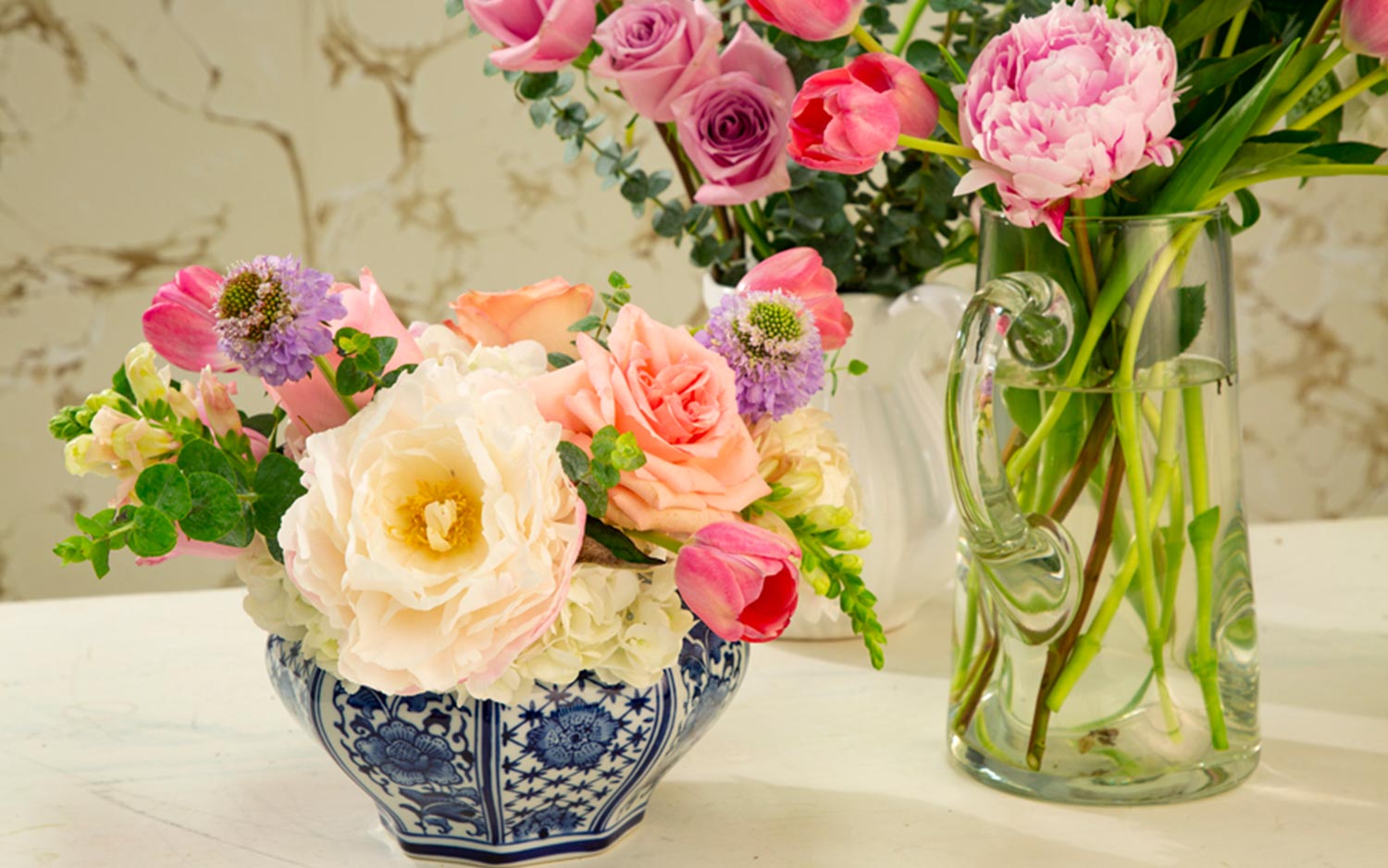 Blue-and-white ceramic vessel and clear glass vase, both full of white, pink, and purple flowers 