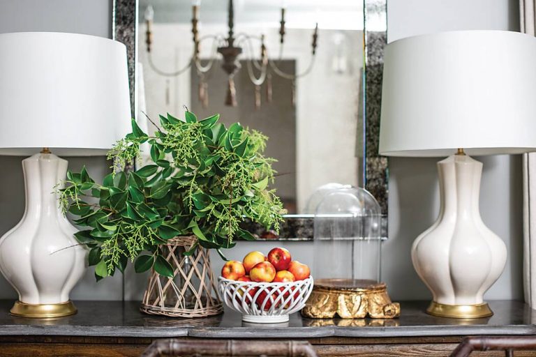 A dining room sideboard vignette comprised of white table lamps, a mirror, a green plant, and a basket of apples