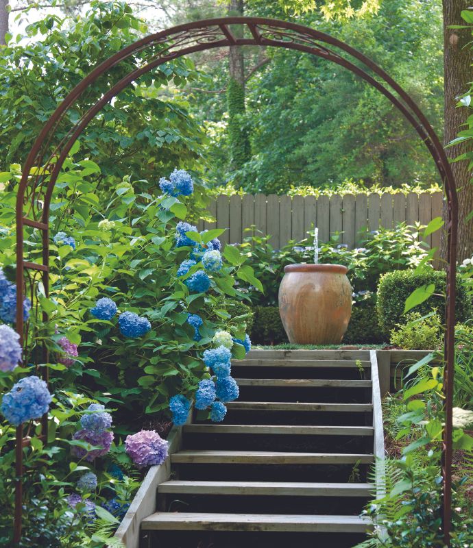 Blue hydrangeas and an iron arch over wooden stairs frames a ceramic fountain