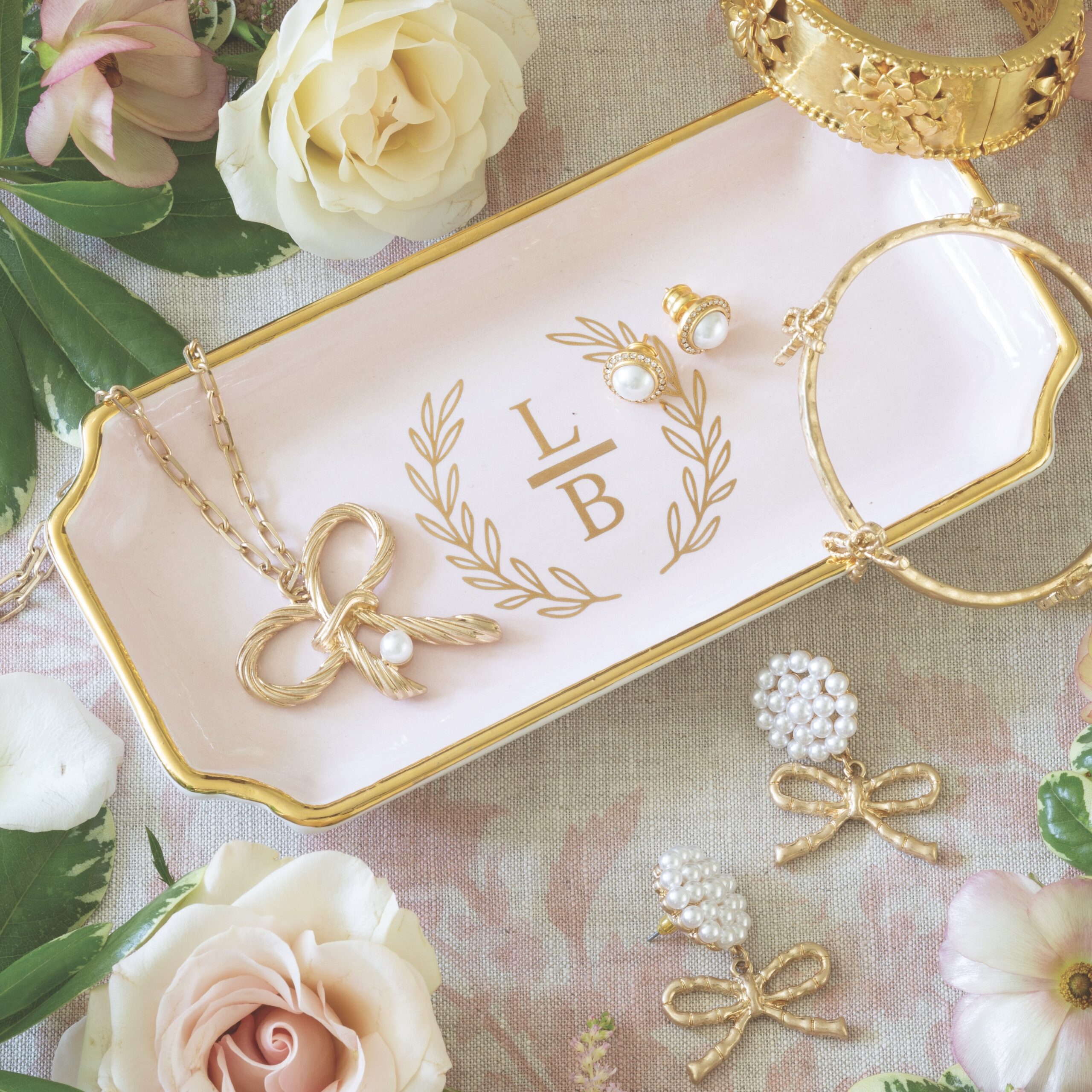 A pink-and-gold monogrammed jewelry tray topped with and surrounded by gold and pearl accessories and flowers