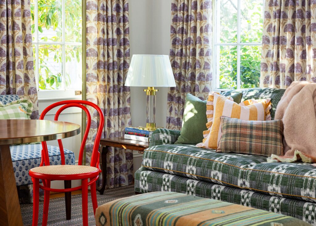 A living room with numerous colorful printed fabrics on the curtains, sofa, pillows, and ottoman, with a red bent wood chair.