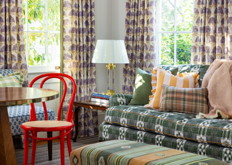 A living room with numerous colorful printed fabrics on the curtains, sofa, pillows, and ottoman, with a red bent wood chair.