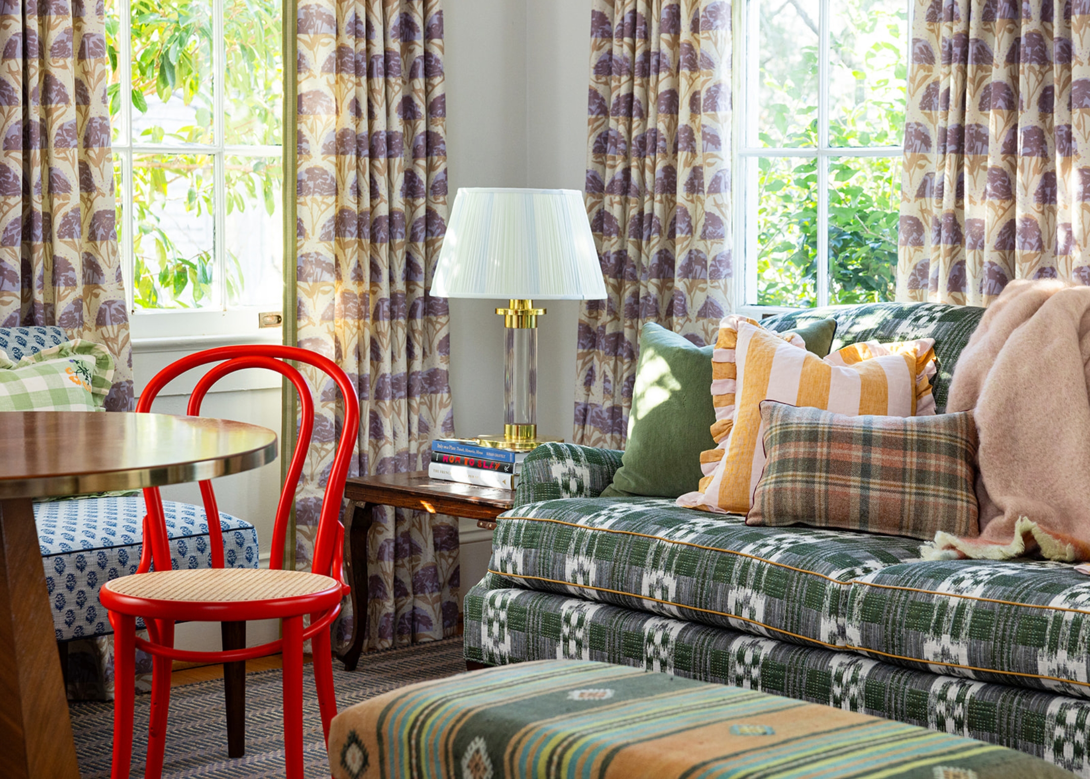 A living room with numerous colorful printed fabrics on the curtains, sofa, pillows, and ottoman, with a red bent wood chair.