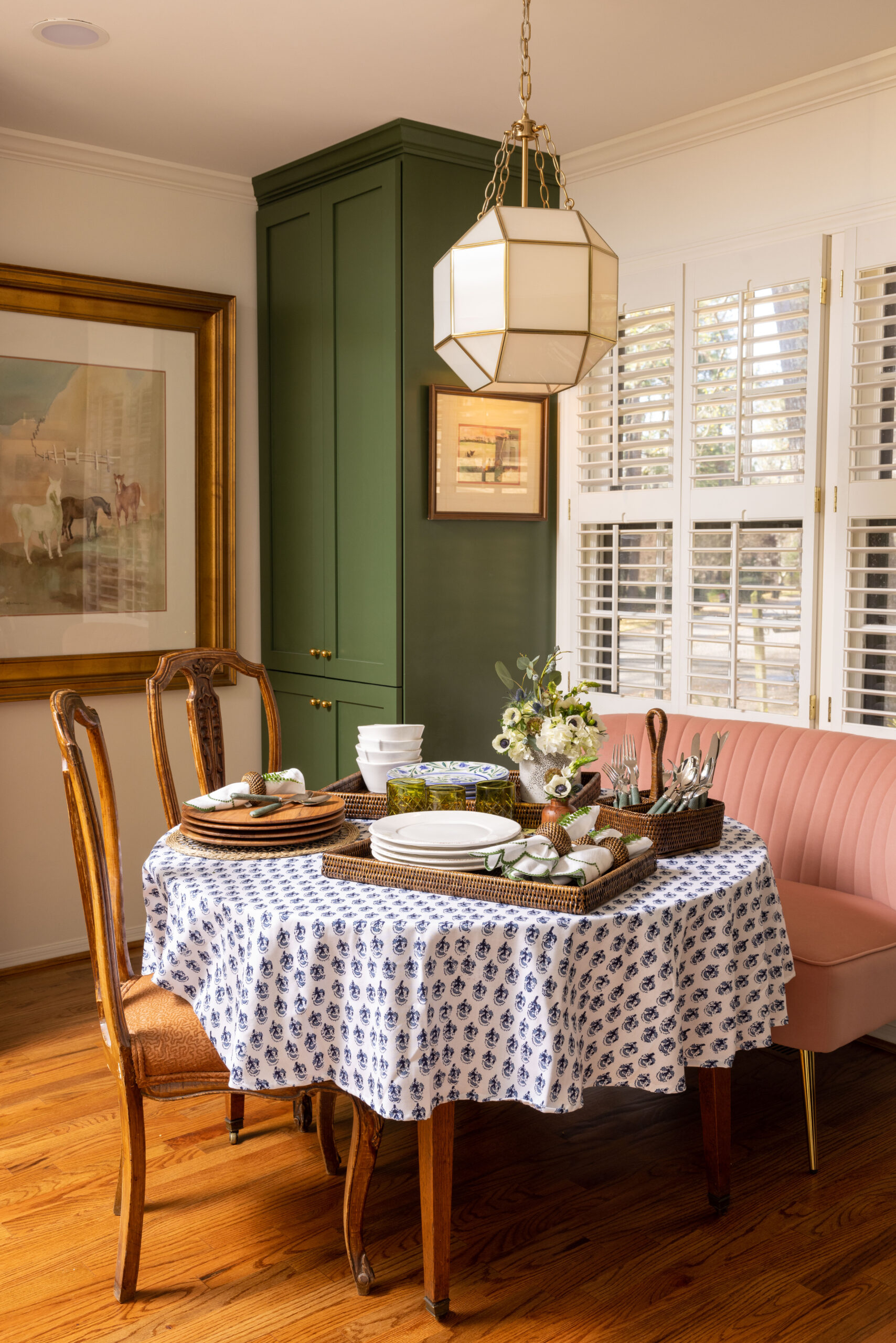 A breakfast nook in a kitchen with green cabinets, traditional artwork, a pink velvet upholstered bench, vintage wooden chairs, a geometric glass pendant light, and a table set with dishware for guests.