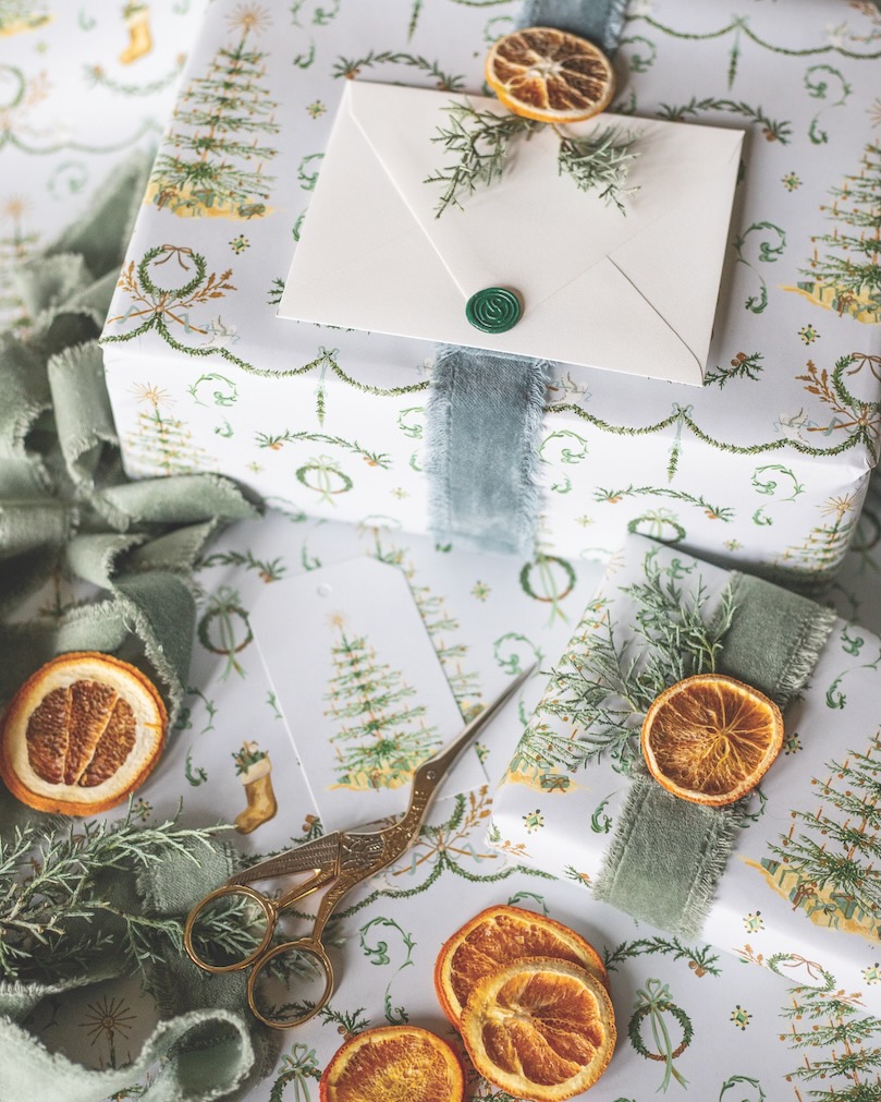 Gifts wrapped with paper printed with Christmas trees, along with ribbons, dried orange slices, and sprigs of greenery.