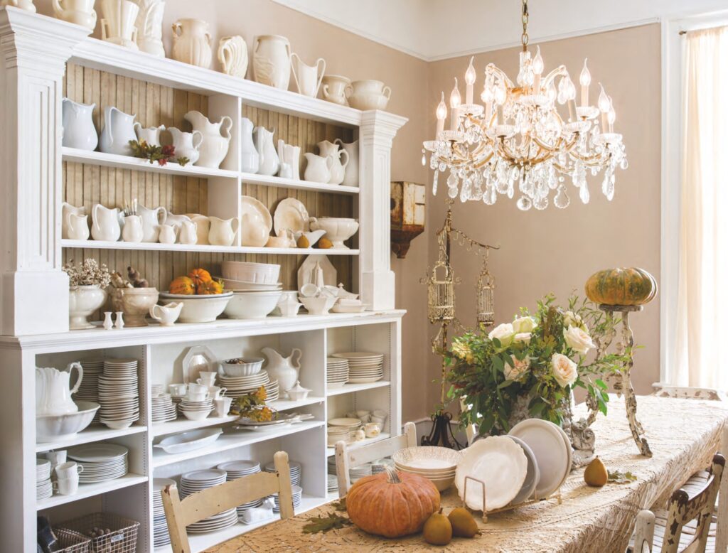 An extensive collection of white ironstone china dishware, including plates, pitchers, cups, and vases, sits on a white hutch in a dining room with a chandelier.