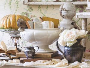 An ironstone pedestal bowl filled with old books, styled with antique silver vases, roses, and pumpkins.