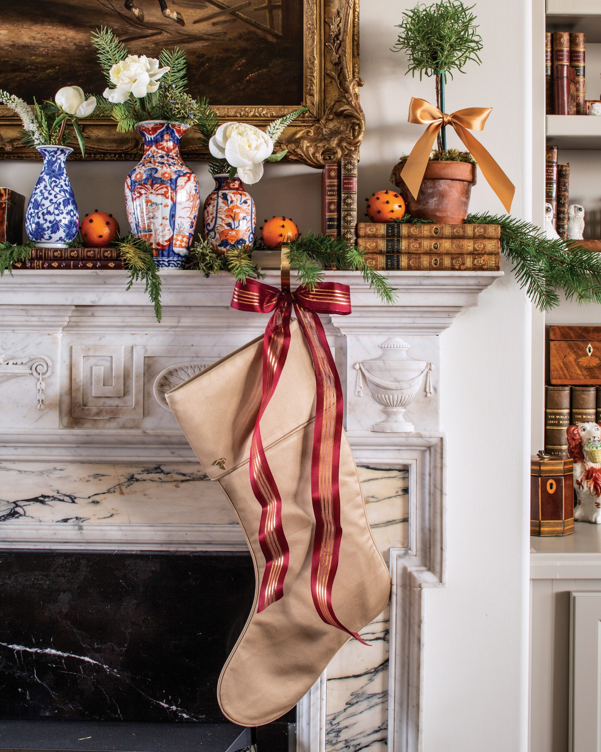A marble fireplace mantel adorned with a stocking, ribbons, chinoiserie vases, tiny topiary trees, books, and pomander oranges.