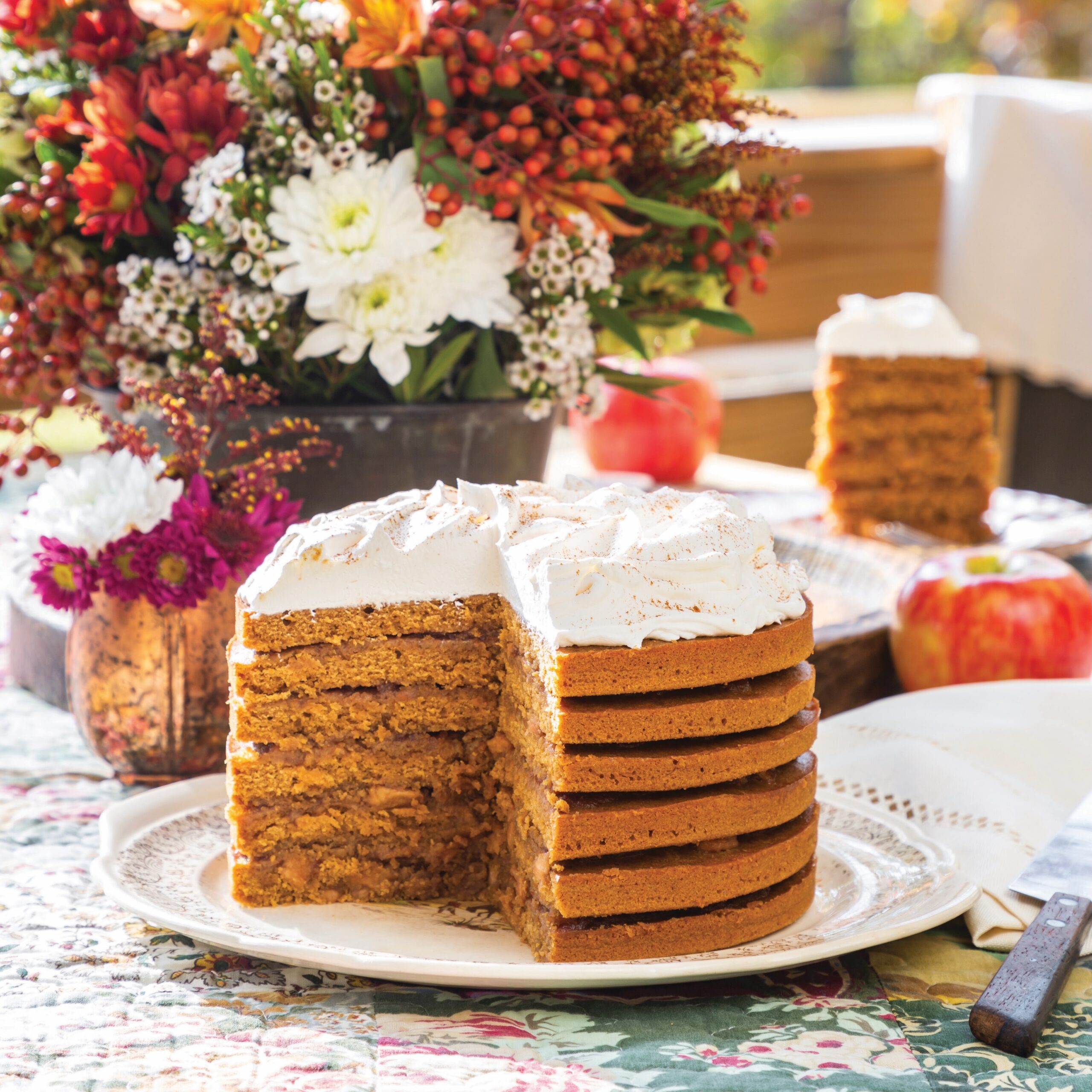 Apple Stack Cake topped with whipped cream and filled with apple butter, on an alfresco table with a fall flower arrangement.