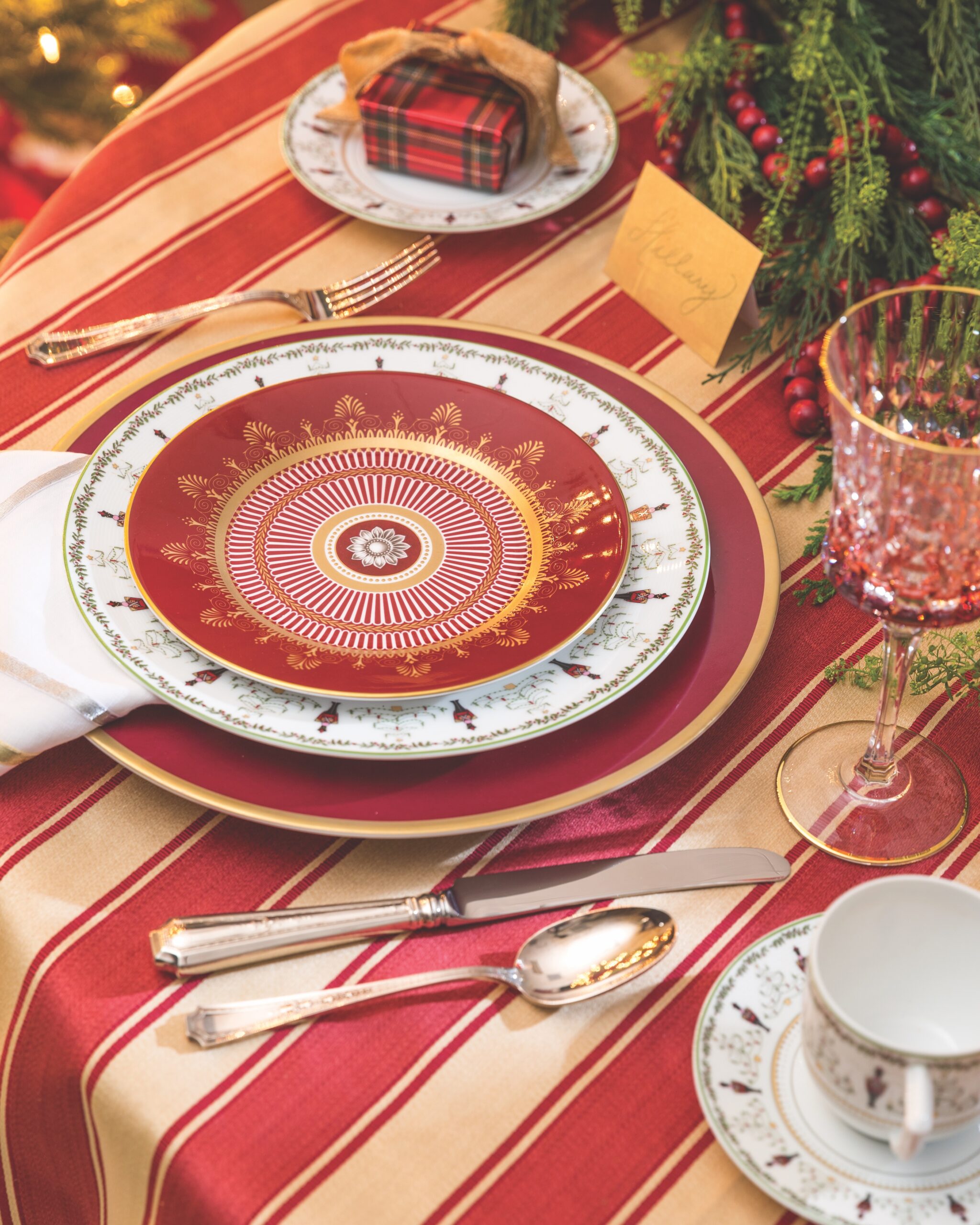 A cranberry-and-gold presentation plate and coordinating striped table linen create the base for a duo of eye-catching china designs. Dainty nutcrackers and artful trees grace Bernardaud's Grenadiers dinner plate and matching service pieces, while a stunning architectural ruby salad dish crowns the setting with a burst of drama. Round out the dynamic display with heirloom silverware and a centerpiece of fresh greenery.