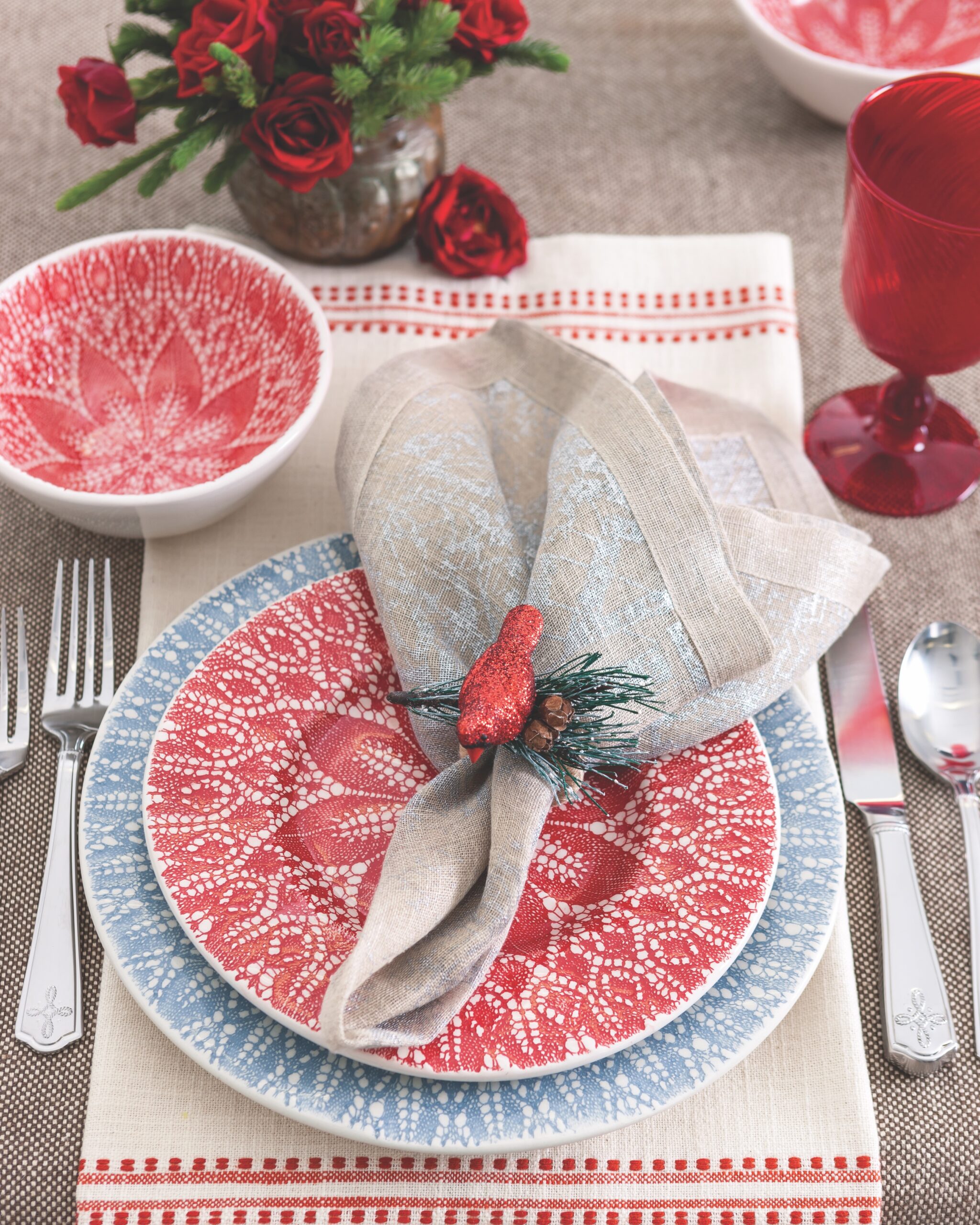 A chipper red bird perched atop a neutral napkin with a subtle silver print creates a sprightly air In this setting. The inspiration plate, Vietri's Lace Gray, is brightened by a matching red holiday version that makes any meal gleeful. Bold glassware and ruby roses complement the china, while the light, intricate pattern of the Villandry Bright Satin flatware by Juliska adds the softness of newly fallen snow.