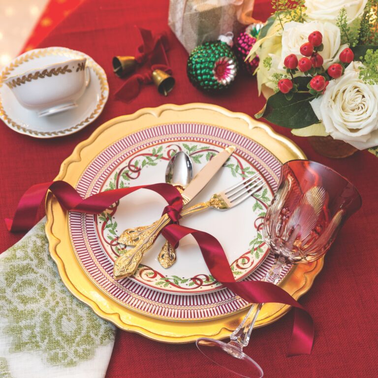 A Christmas place setting consisting of Pickard's Winter Festival salad plate, Wedgwood's Anthemion Ruby dinner plate, a gold charger, and an embroidered napkin with a red tinted goblet.