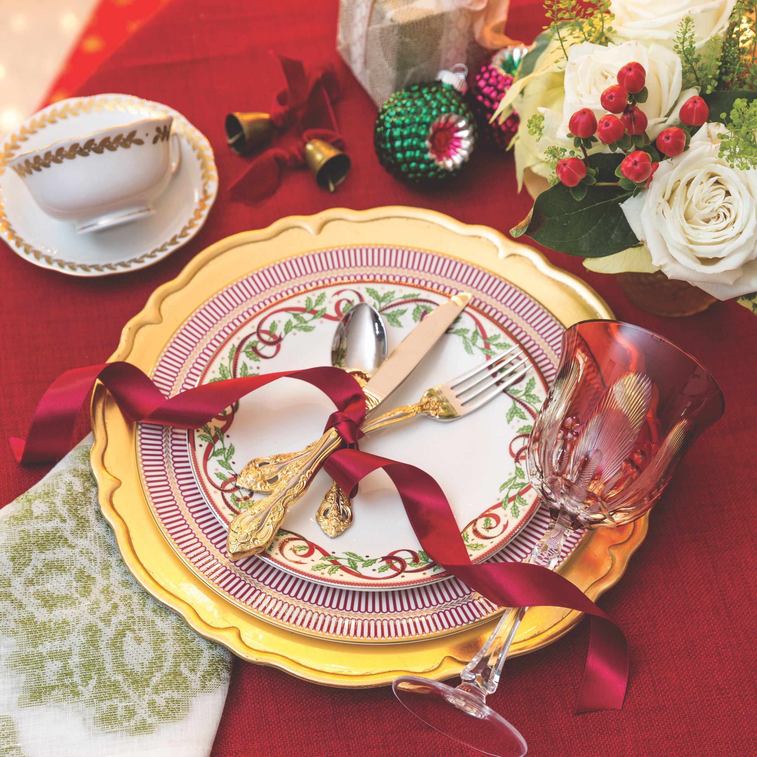 A Christmas place setting consisting of Pickard's Winter Festival salad plate, Wedgwood's Anthemion Ruby dinner plate, a gold charger, and an embroidered napkin with a red tinted goblet.