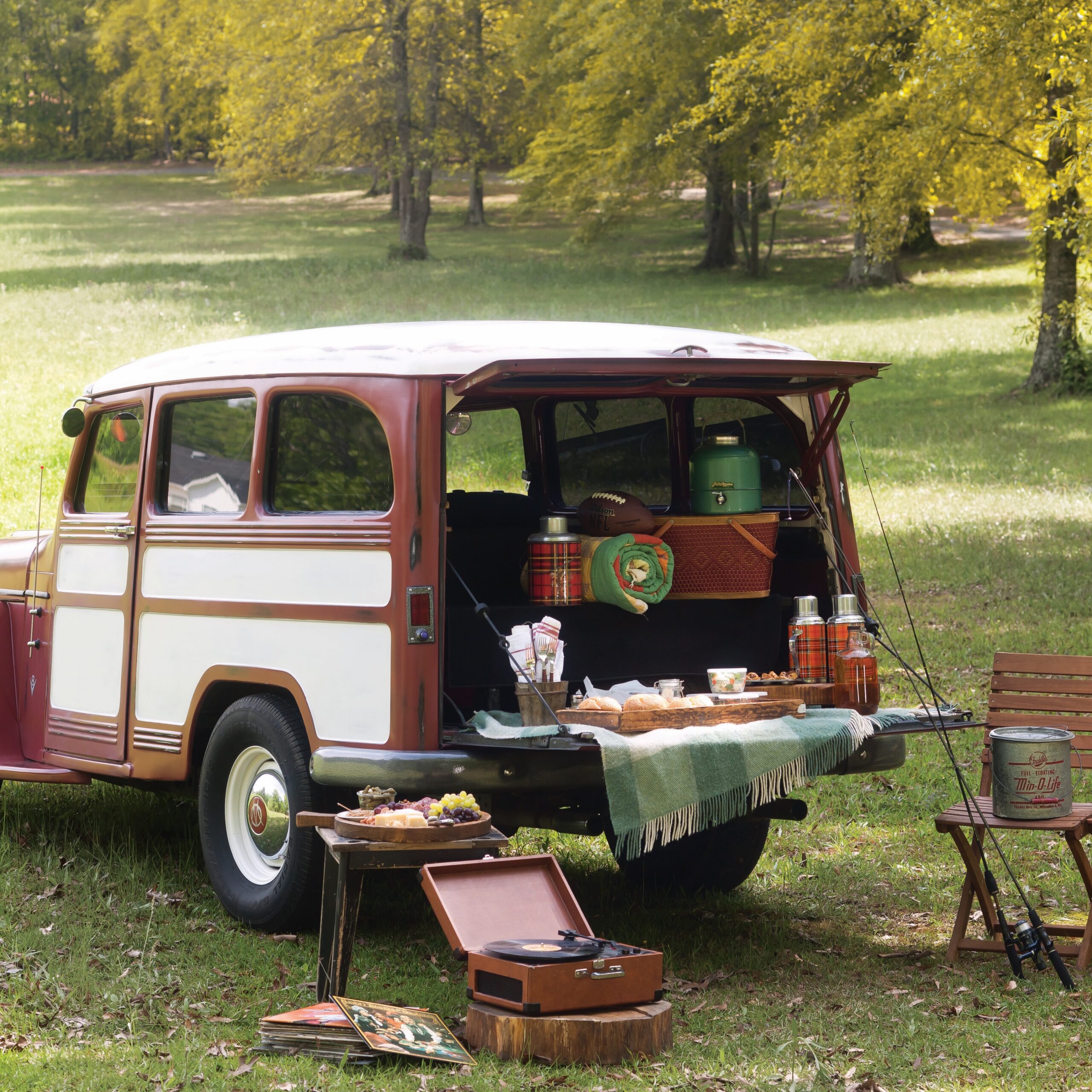 A vintage station wagon with the tailgate down, filled with supplies ofr a picnic, near folding chairs and a record player outdoors.