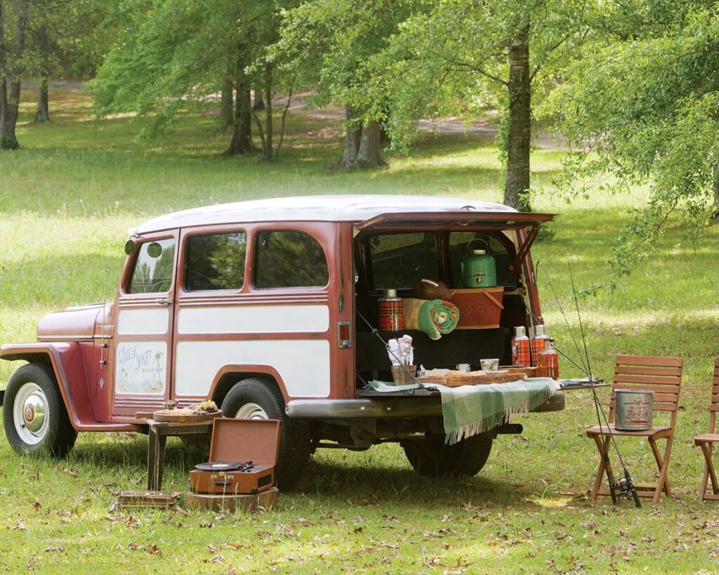 A vintage station wagon with the tailgate down, filled with supplies ofr a picnic, near folding chairs and a record player outdoors.