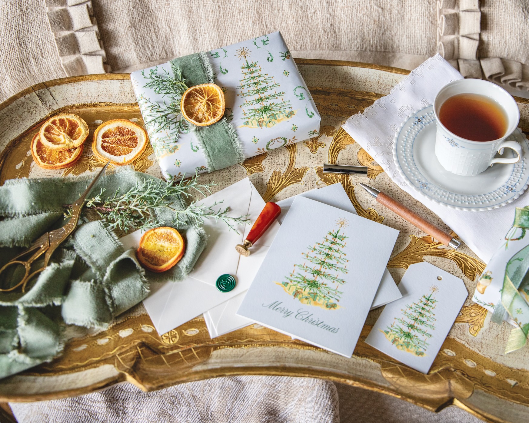 A gift wrapped in the Blissful Holiday gift wrap with a ribbon, sprig, and orange slice, near matching note cards, gift tags, and a cup of tea.