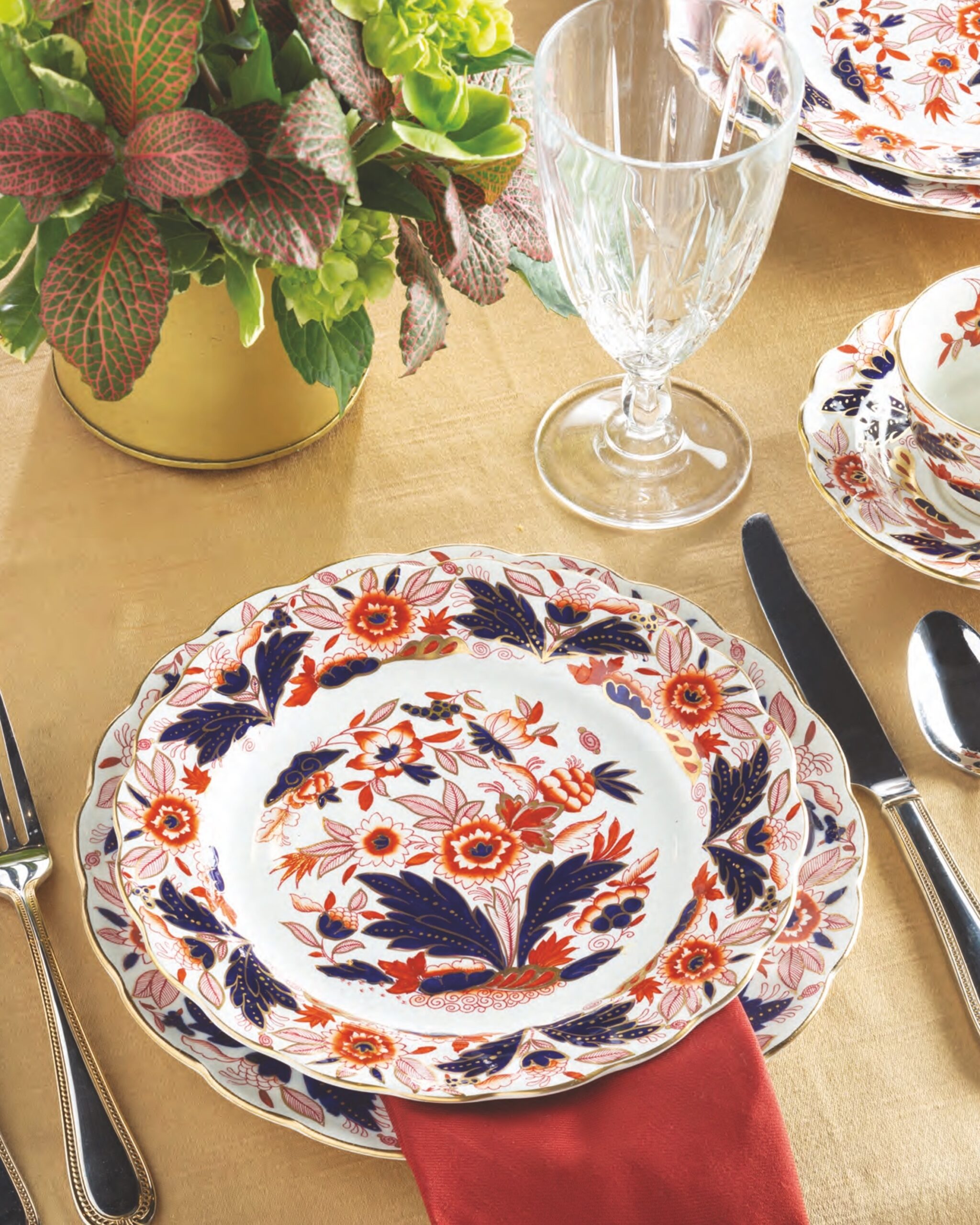 A place setting with Dovedale Rust and Blue with Gold Edge plates, a red napkin, a tan tablecloth, and a crystal goblet.