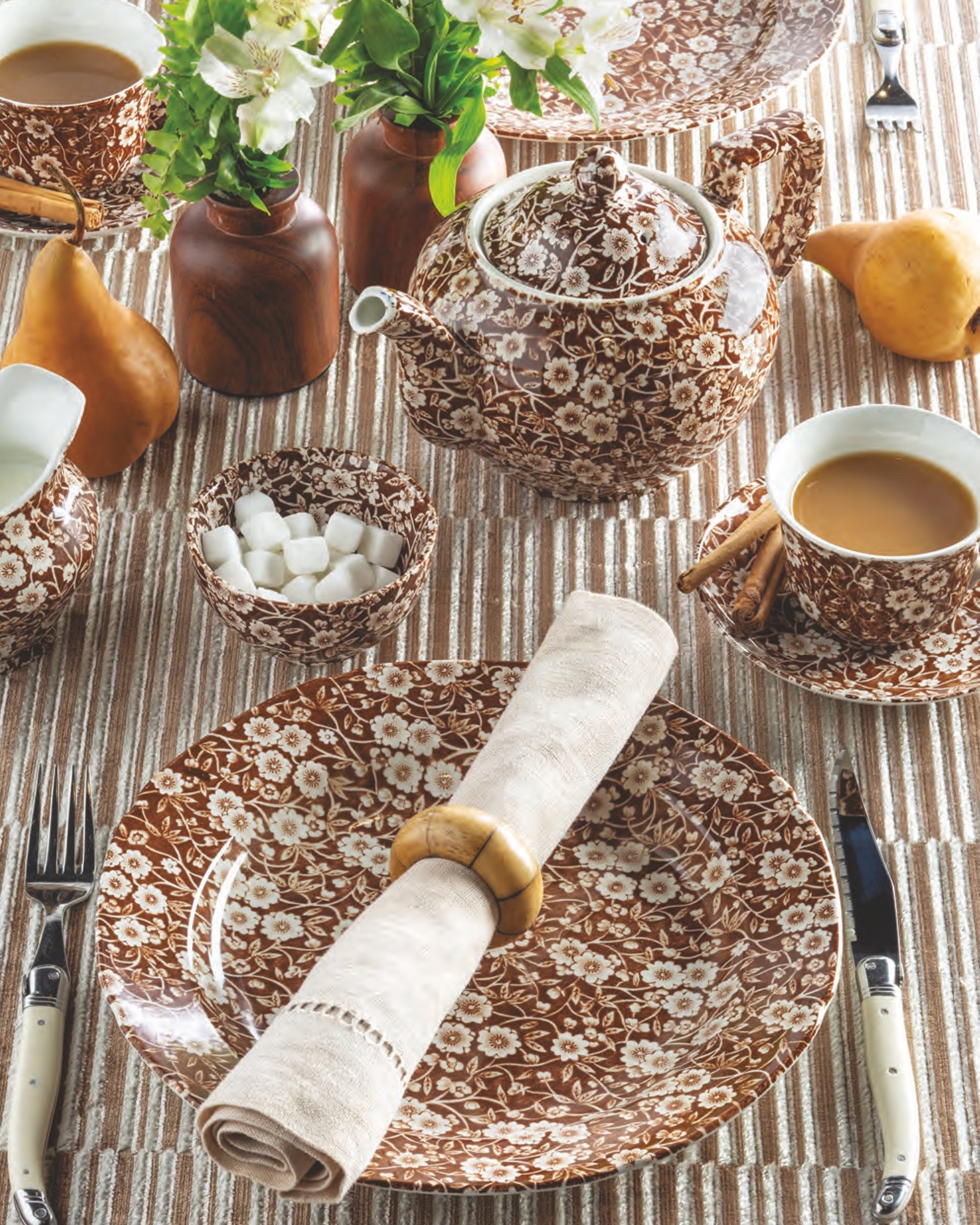 A place setting with the Calico Brown by Staffordshire dinner plate, topped with a linen napkin, with a matching teapot and teacup with saucer on a modern striped tablecloth.