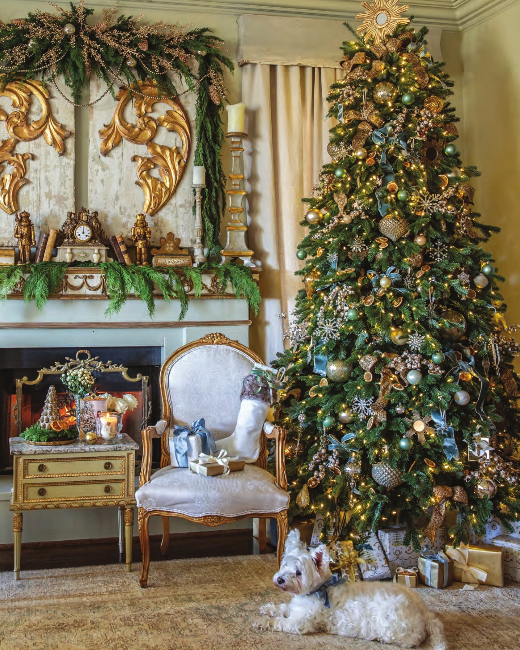 An elaborate French-style living room in a gold, beige, and blue palette with a Christmas tree, mantel draped in garland, terrier, and gifts under the tree