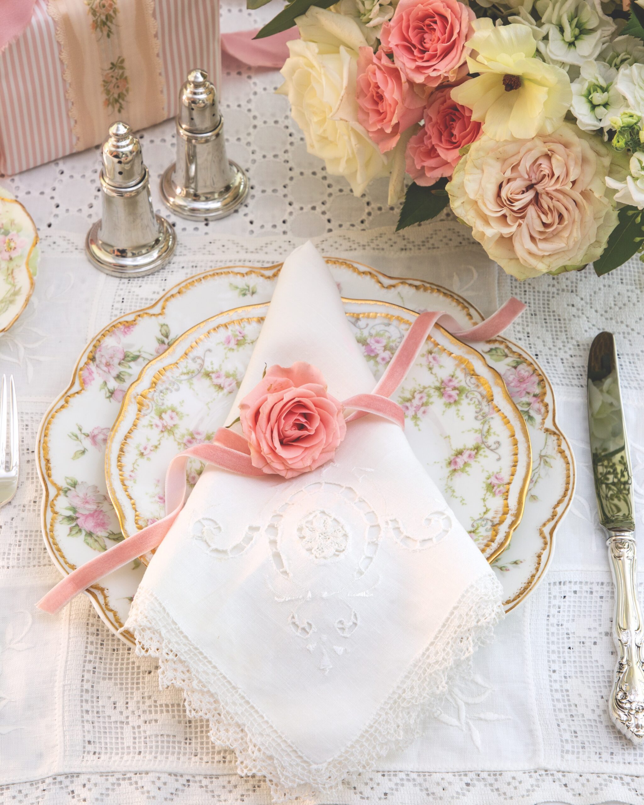 A place setting with floral china, a lace napkin, a rosette, and a pink velvet ribbon near flowers.