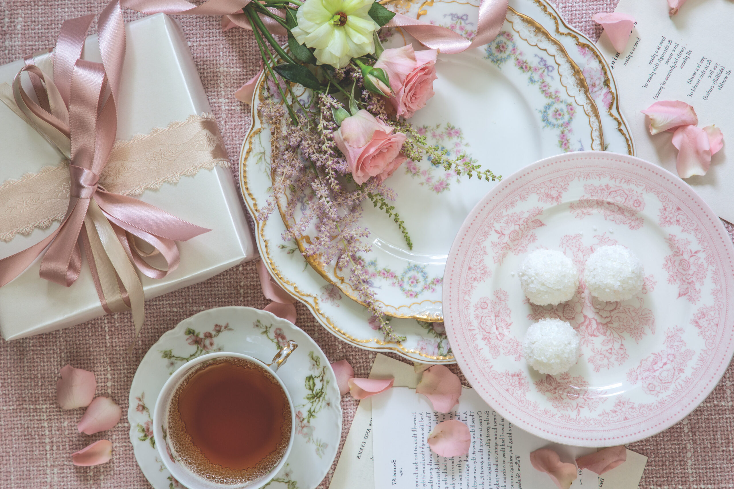 A table scattered with pages of poetry and rose petals is a charming backdrop for collected china united by understated pastel hues. A fetching bouquet tied with a satin sash is the finishing touch to this beguiling tableau, sure to captivate all who gather nearby.
