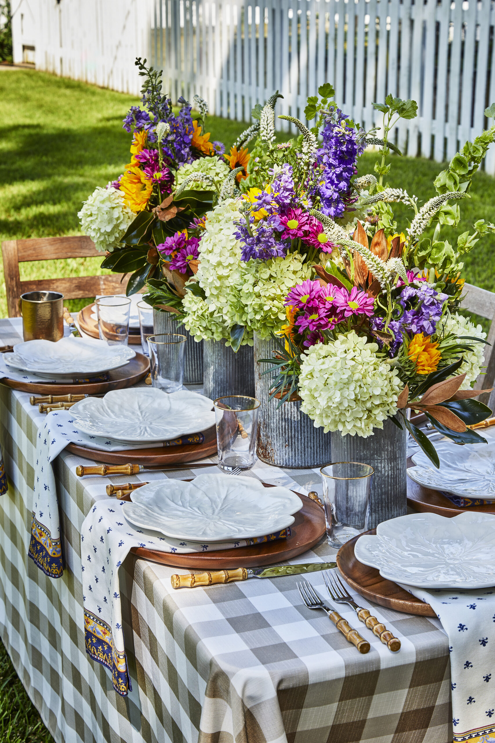 An alfresco table arranged with place settings, colorful flower centerpieces, and a gingham tablecloth.