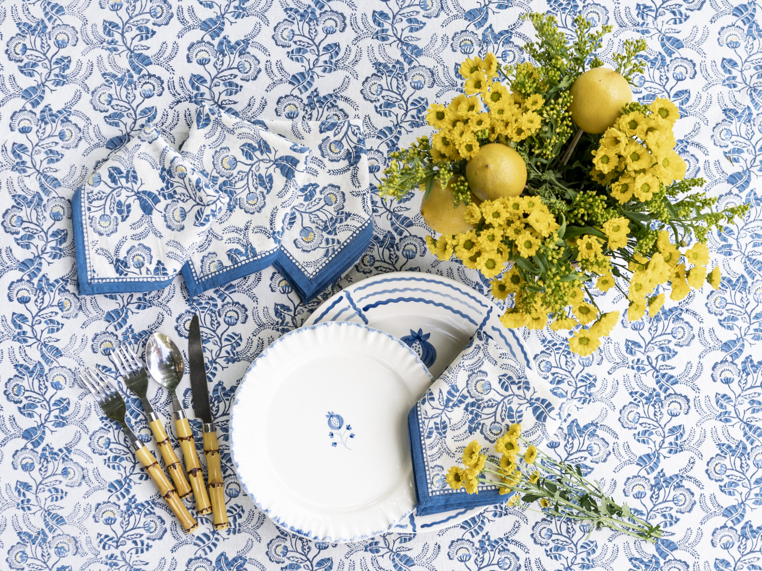 Blue and white floral table linens from Pomegranate arranged in a place setting with a yellow flower arrangement.