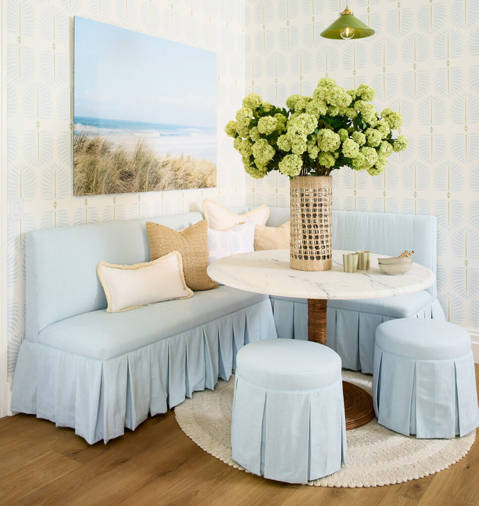 A cozy nook with light blue bench seating, including a pleated skirt, a beachy landscape art piece, and a table with an abundant hydrangea arrangement.