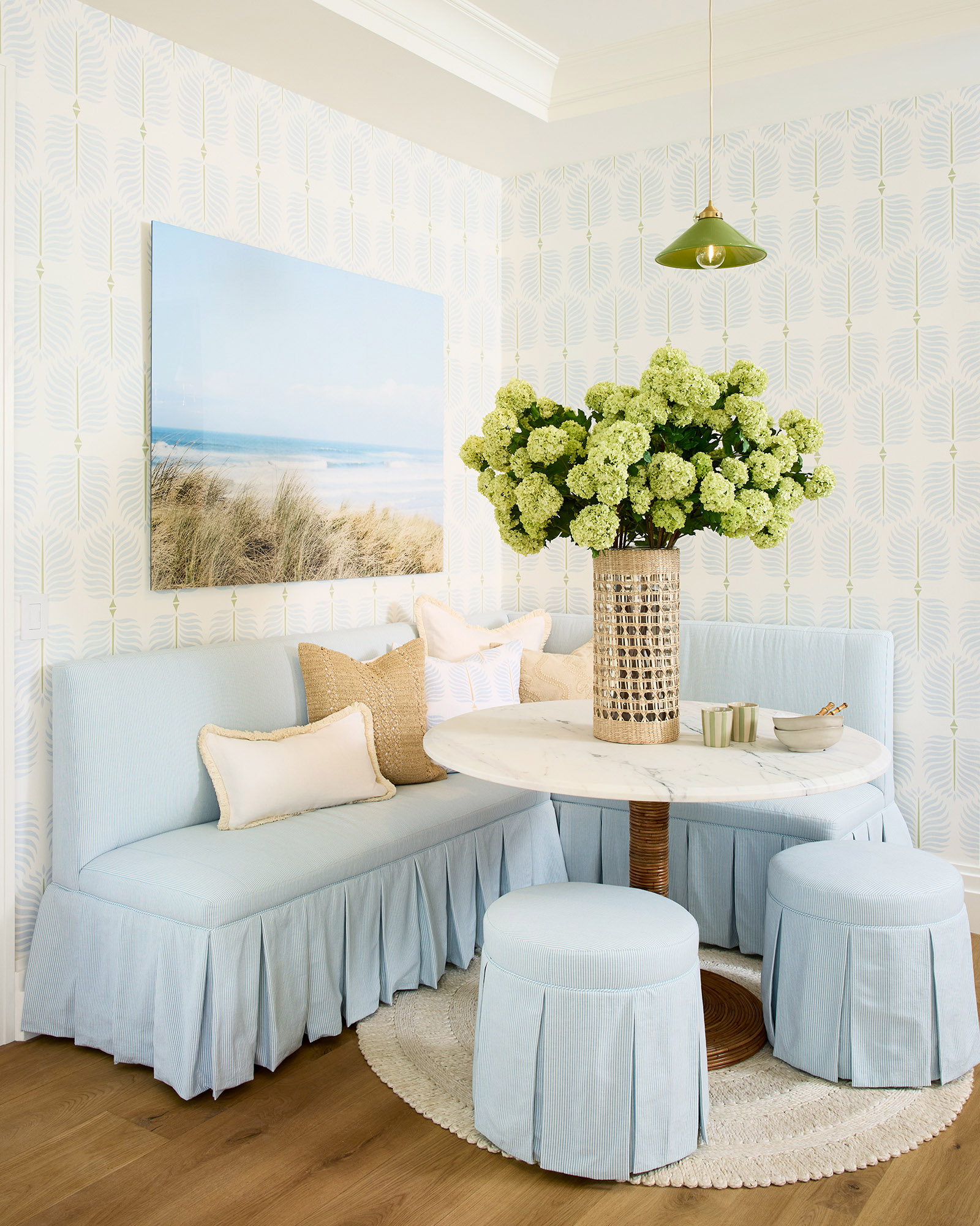 A cozy nook with light blue bench seating, including a pleated skirt, a beachy landscape art piece, and a table with an abundant hydrangea arrangement.