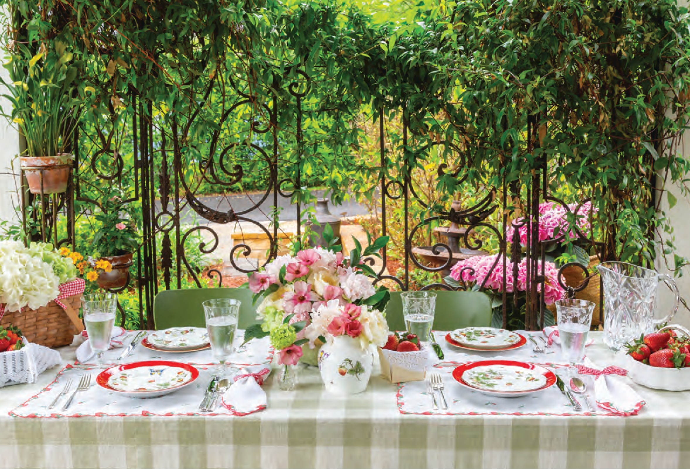 An outdoor table setting with a green gingham tablecloth and strawberry-themed dishware and table decorations, with an abundant flower arrangement.