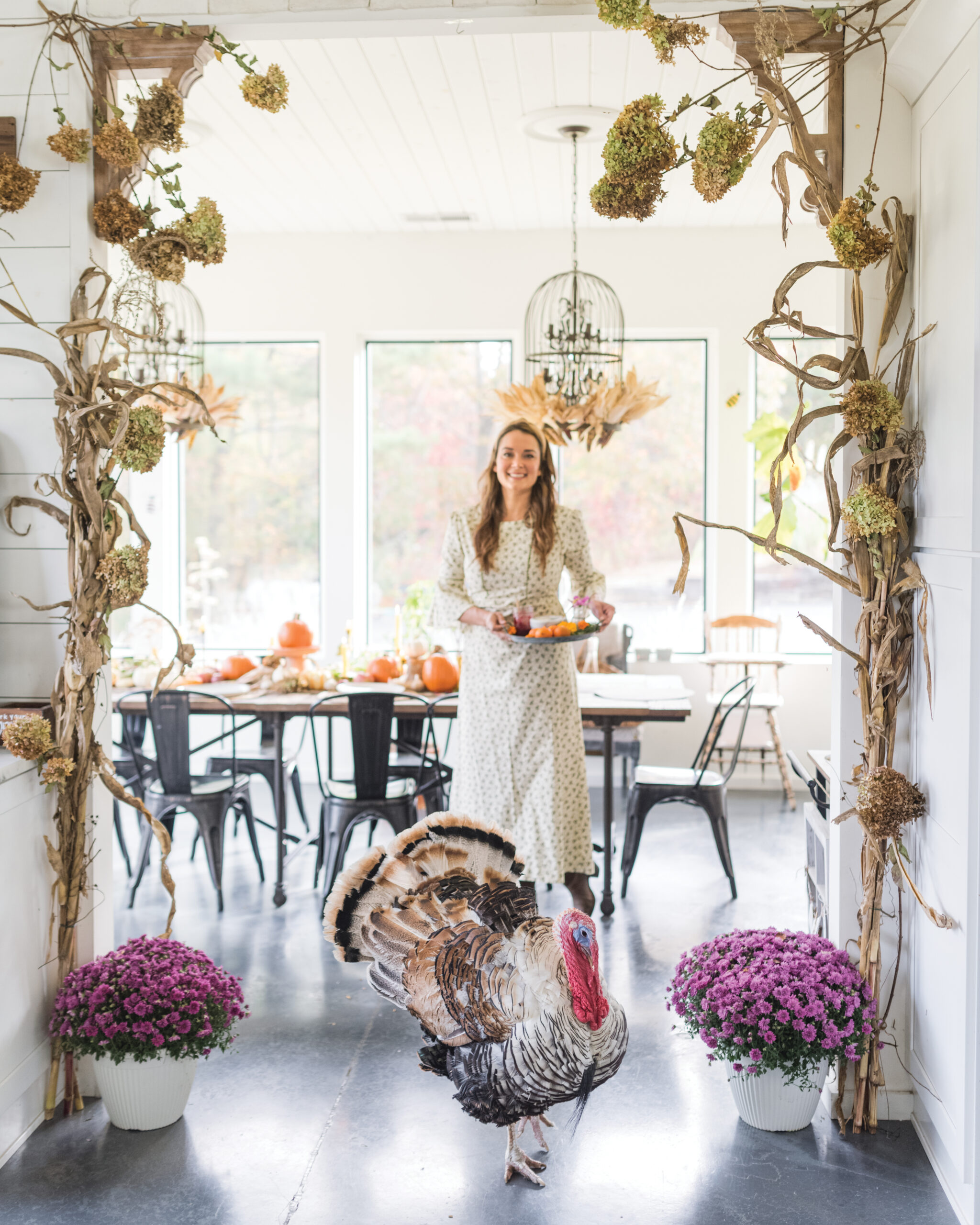 A woman in a light dress carries a tray of food toward a table set with pumpkins, while a turkey walks in the foreground of a festive fall-decorated room.