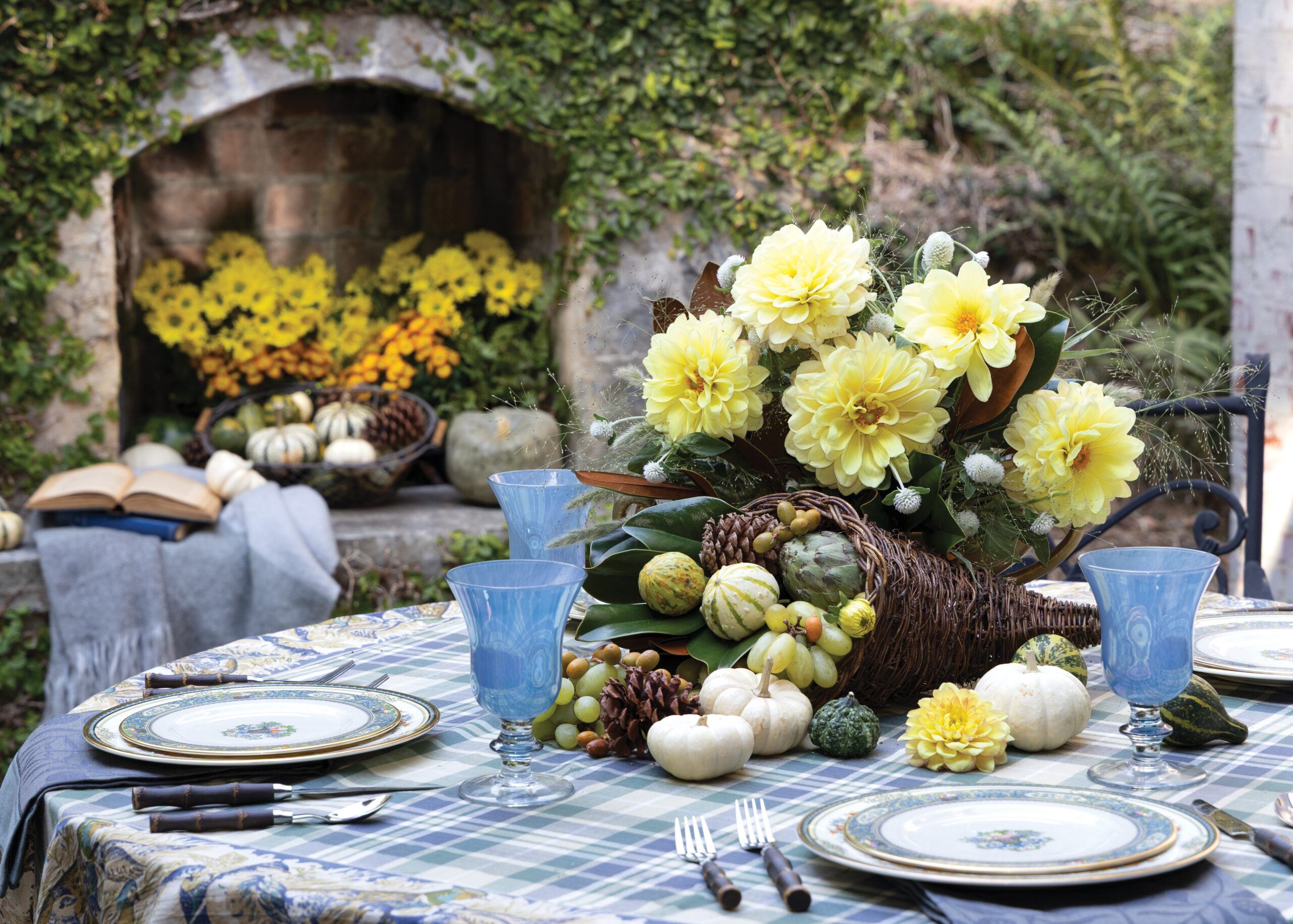A fall-themed outdoor table setting with a cornucopia centerpiece of yellow flowers, gourds, and pumpkins