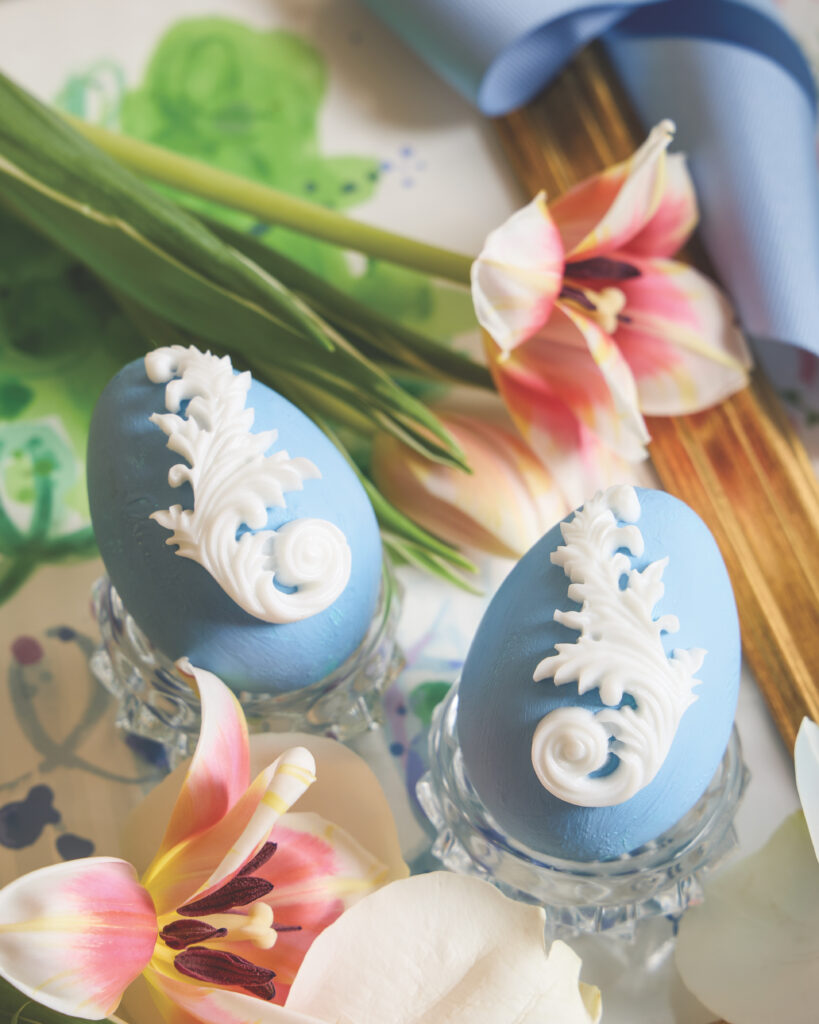 Two blue eggs with a intricate white decal atop it, with pink tulips behind.