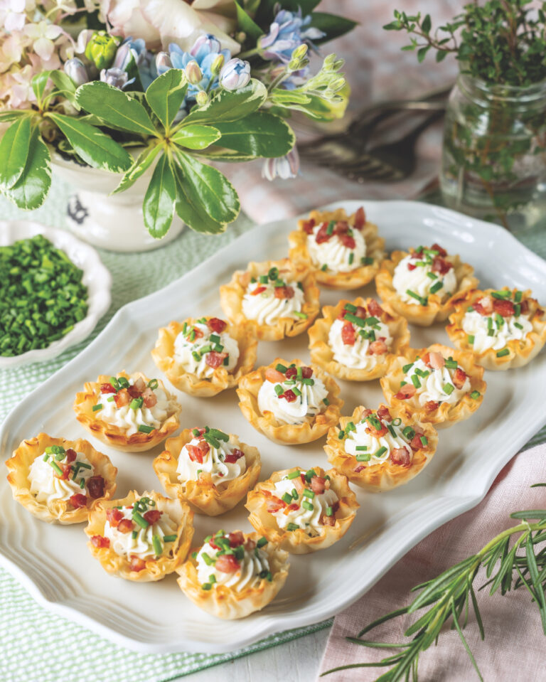 Herb and goat cheese tartlets lined up on a platter surrounded by herbs and flowers.