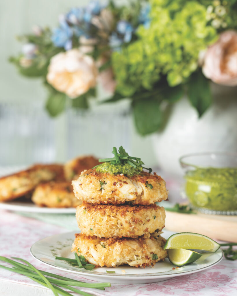 Three crab cakes stacked together with limes on the side, and a green sauce in the background.
