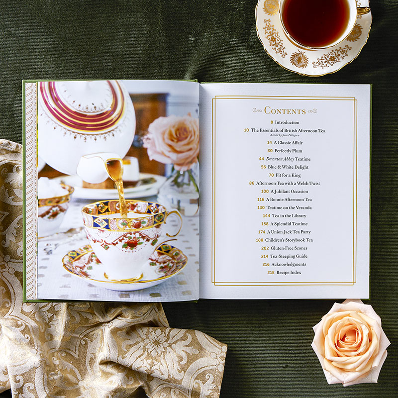 Open cookbook on a green table with a ornate teacup and poured tea on the left page; contents page on the right in gold-frame design.