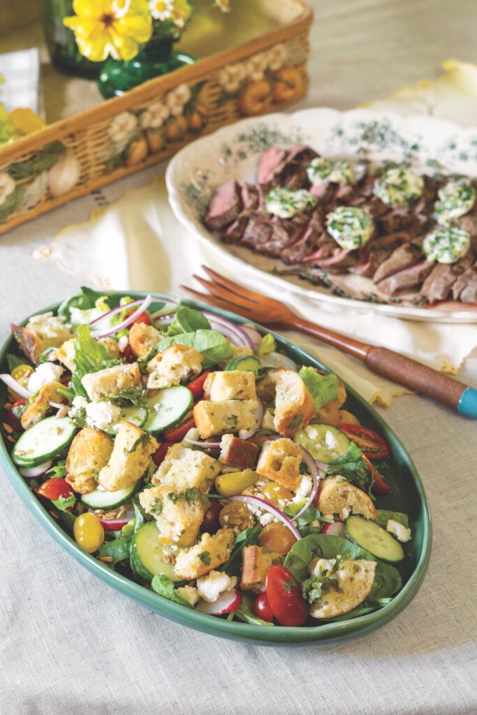 Panzanella salad in an oval serving dish with a platter of steak in the background.