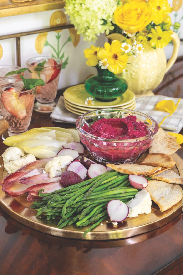 Roasted beet hummus in a clear bowl with various vegetables and crostini served around it on a gold platter.