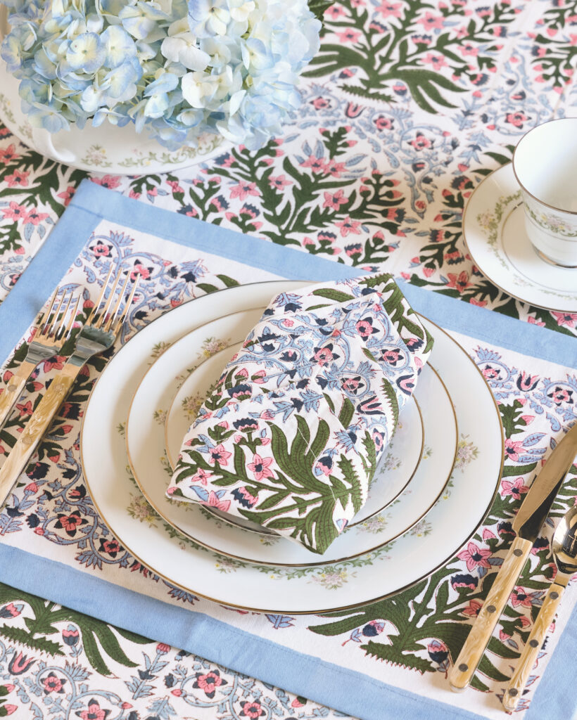 A tablecloth, placemat, and napkin in the same pink, green, and blue block-print pattern, with faux-horn silverware and a trio of dishes.