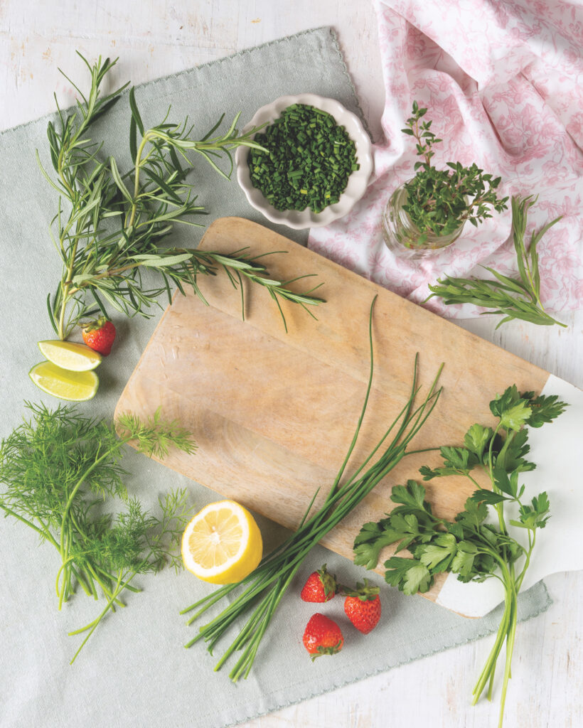 A wooden cutting board surrounded by bunches of herbs like dill, taragon, chives, and parsley with lemons and strawberries.