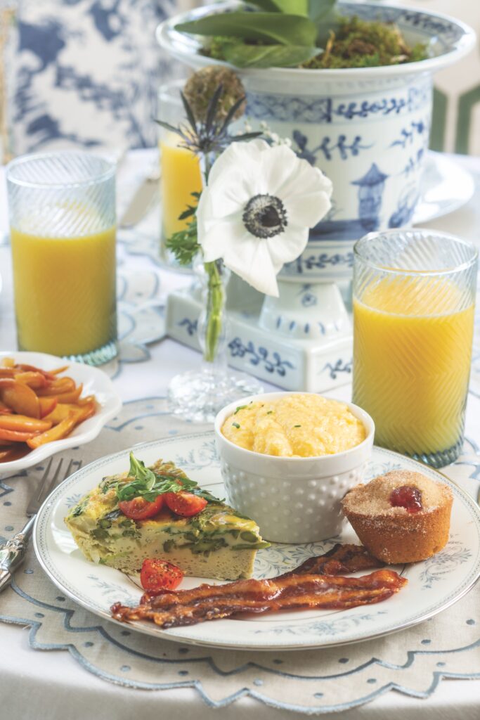 A white plate filled with bacon, frittata, a muffin, and a bowl of cheese grits with a glass of orange juice and cinnamon apples on the side.