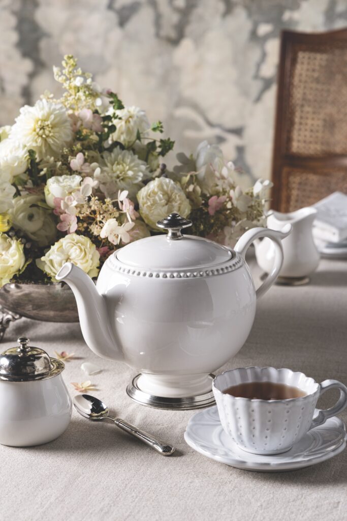 A white teapot with a cup of tea and pot of sugar with a white floral arrangement.