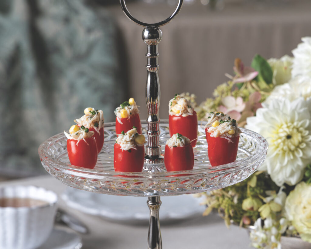 Red stuffed peppers arranged on a glass tiered platter at a catered event with flowers in the background