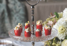 Crab and Corn–Stuffed Cherry Tomatoes Red stuffed peppers arranged on a glass tiered platter at a catered event with flowers in the background