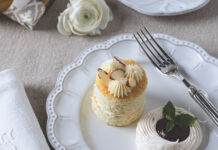 Almond Cakes Two small vanilla cupcakes with white swirled frosting and almond slices on a decorative white plate, with a fork nearby.
