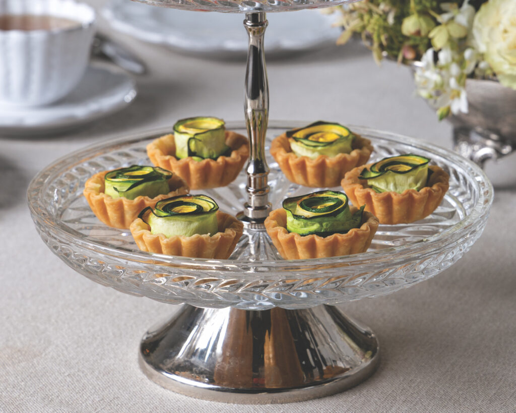 Two-tiered clear glass dessert stand filled with small tartlets topped with curled cucumber ribbons on a light tablecloth