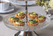 Summer Squash Tartlets Two-tiered clear glass dessert stand filled with small tartlets topped with curled cucumber ribbons on a light tablecloth
