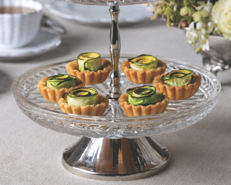 Two-tiered clear glass dessert stand filled with small tartlets topped with curled cucumber ribbons on a light tablecloth