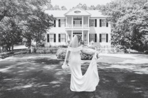 A black-and-white photo of a bride standing in front of a white manor.